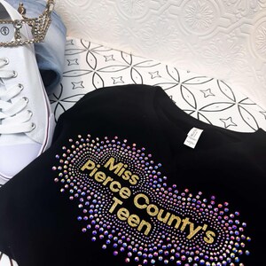 Puede incluir: Camiseta negra con un dise&ntilde;o de purpurina dorada que dice "Miss Pierce County's Teen" rodeado de pedrer&iacute;a iridiscente.