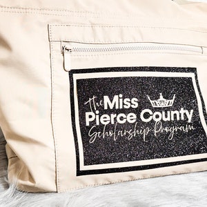 May include: A beige tote bag with a zippered pocket and a black and white rectangular patch. The patch features the text "The Miss Pierce County Scholarship Program" with a crown graphic. The bag is made of a smooth, light-colored material.