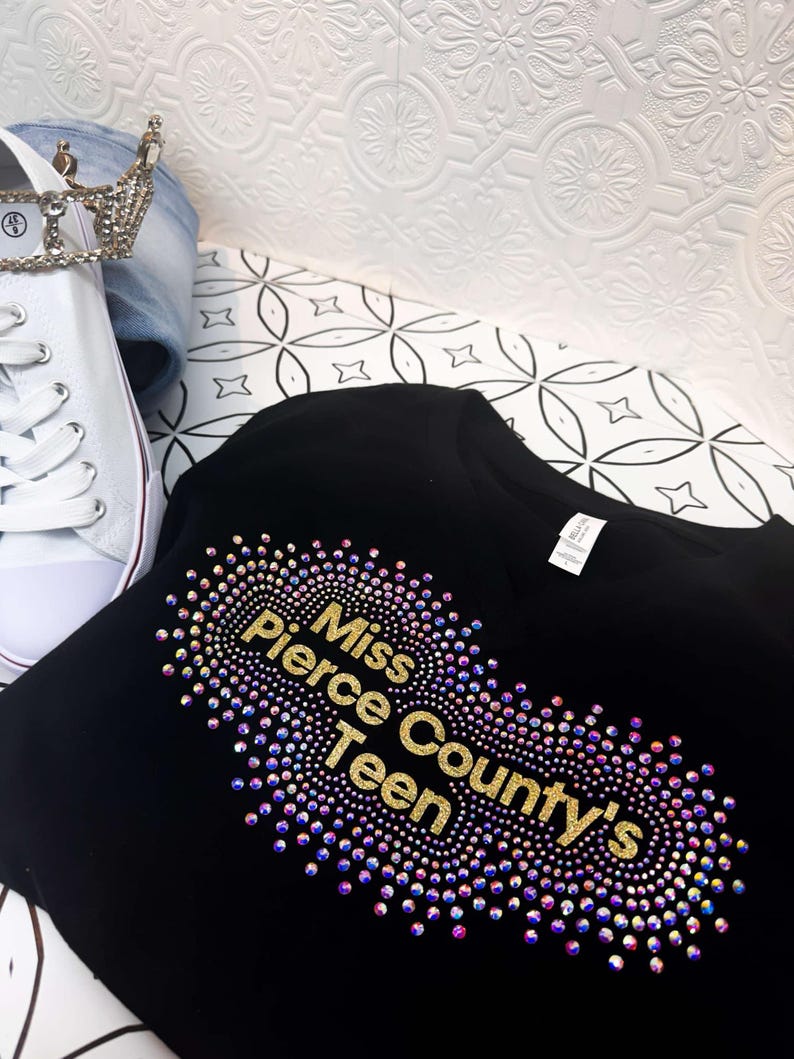 Puede incluir: Camiseta negra con un dise&ntilde;o de purpurina dorada que dice "Miss Pierce County's Teen" rodeado de pedrer&iacute;a iridiscente.