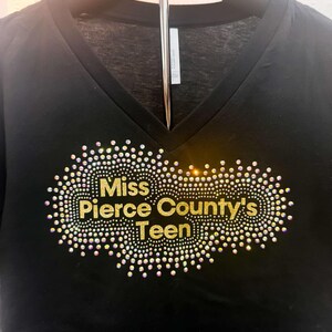 Puede incluir: Camiseta negra de cuello en V con un dise&ntilde;o de purpurina dorada que dice "Miss Pierce County's Teen".