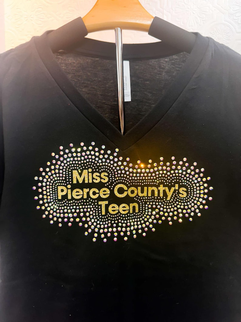Puede incluir: Camiseta negra de cuello en V con un dise&ntilde;o de purpurina dorada que dice "Miss Pierce County's Teen".