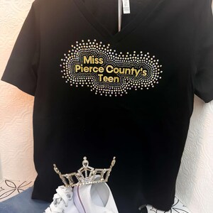 Puede incluir: Camiseta negra de cuello en V con un dise&ntilde;o de pedrer&iacute;a dorada que dice "Miss Pierce County's Teen". Tambi&eacute;n se ve una corona blanca y zapatillas blancas en la imagen.