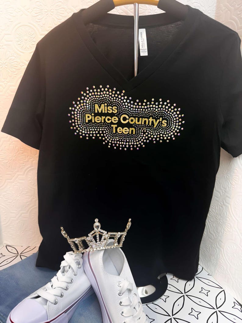 Puede incluir: Camiseta negra de cuello en V con un dise&ntilde;o de pedrer&iacute;a dorada que dice "Miss Pierce County's Teen". Tambi&eacute;n se ve una corona blanca y zapatillas blancas en la imagen.