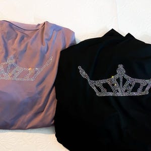 May include: Two tops, one lavender and one black, each adorned with a rhinestone crown design. The tops are folded and rest on a white surface, showcasing the sparkling embellishments.