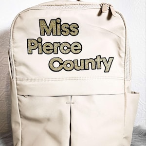 May include: A beige backpack with a zipper closure and a large pocket on the front. The backpack has the text "Miss Pierce County" in gold glitter letters.