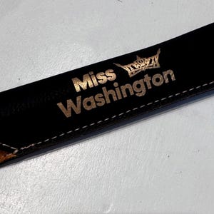 May include: Black leather pen case with the words "Miss Washington" and a crown design in gold lettering. The case has a tan interior and white stitching along the edges. A stylish accessory for pens and pencils.
