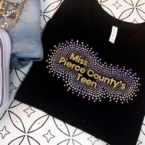 Puede incluir: Camiseta negra con un dise&ntilde;o de purpurina dorada que dice "Miss Pierce County's Teen".