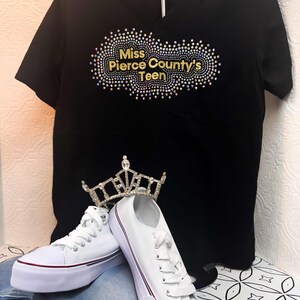 Puede incluir: Camiseta negra de cuello en V con un dise&ntilde;o de purpurina dorada que dice "Miss Pierce County's Teen". La camiseta est&aacute; en una percha y hay zapatillas blancas y una corona en el fondo.