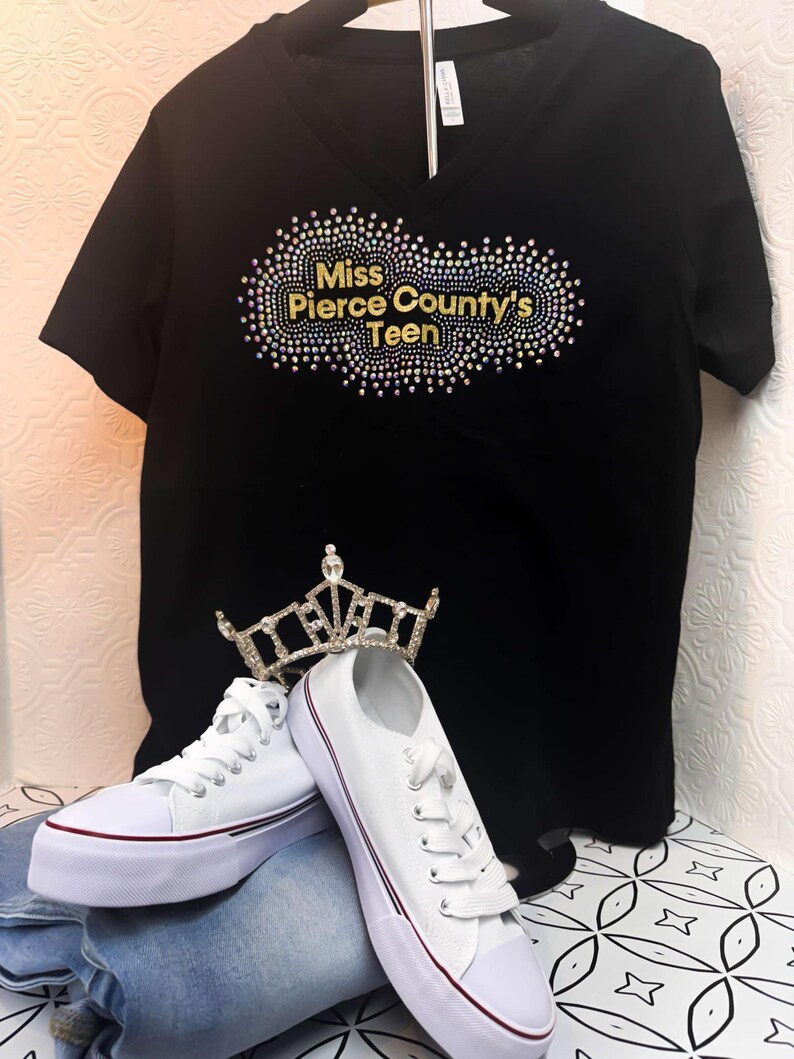 Puede incluir: Camiseta negra de cuello en V con un dise&ntilde;o de purpurina dorada que dice "Miss Pierce County's Teen". La camiseta est&aacute; en una percha y hay zapatillas blancas y una corona en el fondo.
