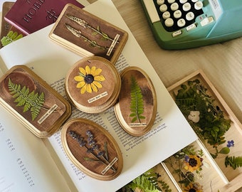 May include: A collection of six oval-shaped wooden frames with pressed flowers and leaves. The frames are arranged on a white surface with a book and a vintage typewriter. The frames feature various botanical specimens, including ferns, lavender, and wildflowers. The frames have handwritten labels with the names of the plants.