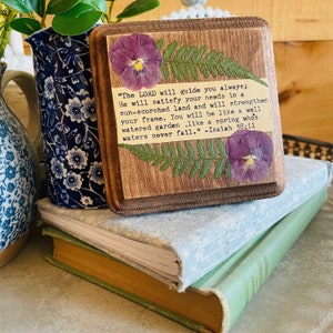 May include: A wooden sign with a pressed flower design and a quote from Isaiah 58:11. The quote reads, "The LORD will guide you always; He will satisfy your needs in a sun-scorched land and will strengthen your frame. You will be like a well-watered garden, like a spring whose waters never fail." -Isaiah 58:11