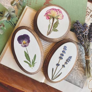 May include: Three oval wooden frames with white backgrounds and pressed flowers. The frames are arranged on a book with green pages. The flowers are a purple pansy, a lavender sprig, and a pink and yellow rose.
