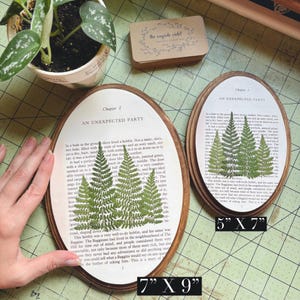 May include: Two oval wooden framed art pieces. The larger one is 7" x 9" and the smaller one is 5" x 7". Each features a page from a book with pressed green ferns. The text "Chapter 1 AN UNEXPECTED PARTY" is visible.
