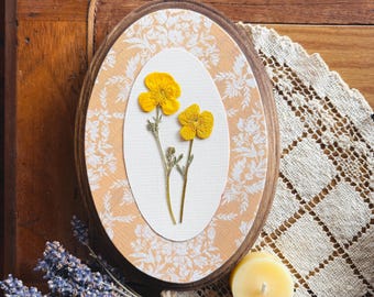 May include: An oval wooden frame with a floral design, featuring two yellow pressed flowers on a white background. The frame is set against a patterned peach background and a wooden surface. A bundle of lavender and a candle are also visible.