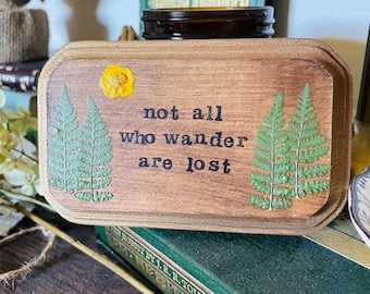 May include: A wooden sign with the quote "not all who wander are lost" printed in black. The sign is decorated with pressed green ferns and a yellow flower.