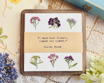 May include: A framed canvas with a quote by Claude Monet: "I must have flowers, always and always." The quote is surrounded by pressed flowers in shades of purple, pink, and white.