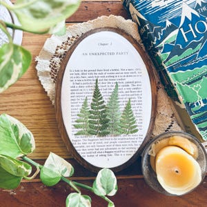 May include: An oval wooden frame with a pressed fern design over a page from a book. The text "Chapter 1 AN UNEXPECTED PARTY" is visible. A copy of "The Hobbit" book is in the background, along with a lit candle.