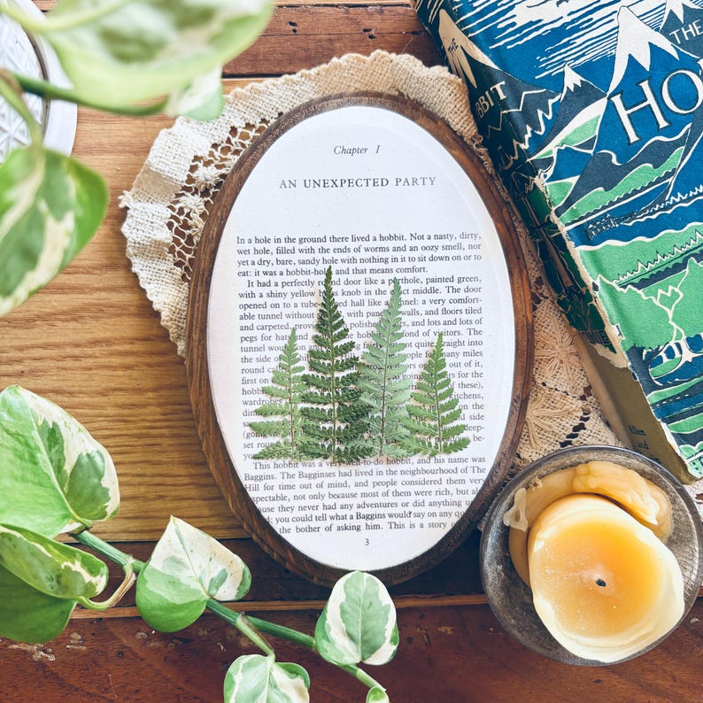 May include: An oval wooden frame with a pressed fern design over a page from a book. The text "Chapter 1 AN UNEXPECTED PARTY" is visible. A copy of "The Hobbit" book is in the background, along with a lit candle.