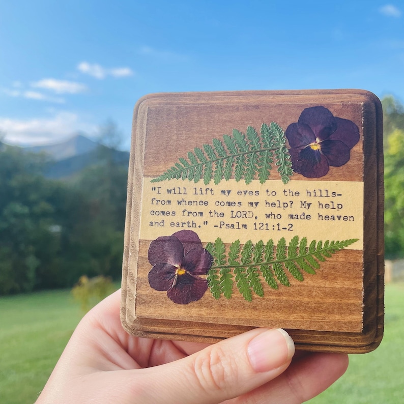 Op de afbeelding: Houten plaquette met een varen en paarse bloemen die in het oppervlak zijn geperst. De plaquette heeft een citaat uit Psalm 121:1-2: "Ik sla mijn ogen op naar de bergen: waar zal mijn hulp vandaan komen? Mijn hulp komt van de HEERE, die hemel en aarde gemaakt heeft." - Psalm 121:1-2