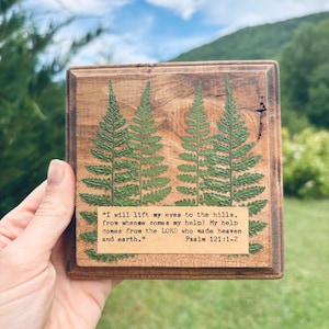 May include: A square wooden plaque featuring two pressed green fern fronds and a rectangular paper insert printed with the text: "I will lift my eyes to the hills, from whence comes my help? My help comes from the LORD who made heaven and earth." Psalm 121:1-2.