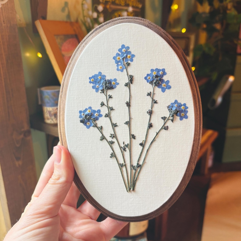 May include: An oval-shaped wall hanging featuring pressed blue forget-me-not flowers arranged on a cream-colored fabric background. The floral arrangement is encased within a dark brown wooden frame with a smooth, rounded edge and a clean finish.