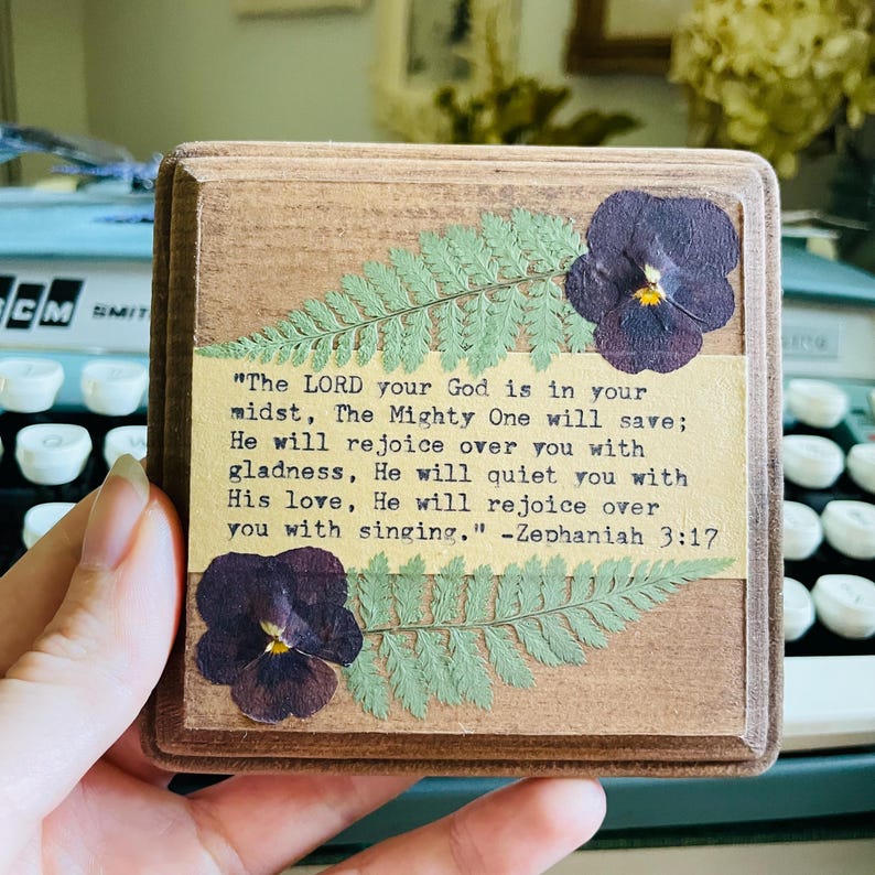 Op de afbeelding: Een houten plaquette met een ontwerp van geperste bloemen en een bijbelvers uit Zefanja 3:17. De tekst luidt: "De HEERE, uw God, is in uw midden, de Machtige zal redden; Hij zal zich over u verheugen met blijdschap, Hij zal u met Zijn liefde sussen, Hij zal zich over u verheugen met zang."