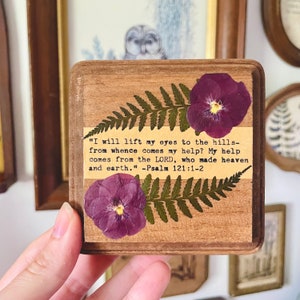 Op de afbeelding: Een houten plaquette met een geperste varen en paarse viooltjes. De plaquette heeft een citaat uit Psalm 121:1-2: "Ik sla mijn ogen op naar de bergen: waar zal mijn hulp vandaan komen? Mijn hulp komt van de HEERE, die hemel en aarde gemaakt heeft." - Psalm 121:1-2