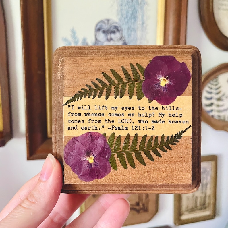 Op de afbeelding: Een houten plaquette met een geperste varen en paarse viooltjes. De plaquette heeft een citaat uit Psalm 121:1-2: "Ik sla mijn ogen op naar de bergen: waar zal mijn hulp vandaan komen? Mijn hulp komt van de HEERE, die hemel en aarde gemaakt heeft." - Psalm 121:1-2
