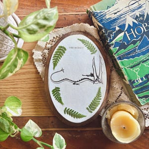 May include: Oval wooden plaque with a white background, featuring a black dragon illustration and the text "THE HOBBIT." Green fern leaves frame the design. A vintage book with a mountain landscape cover is visible.