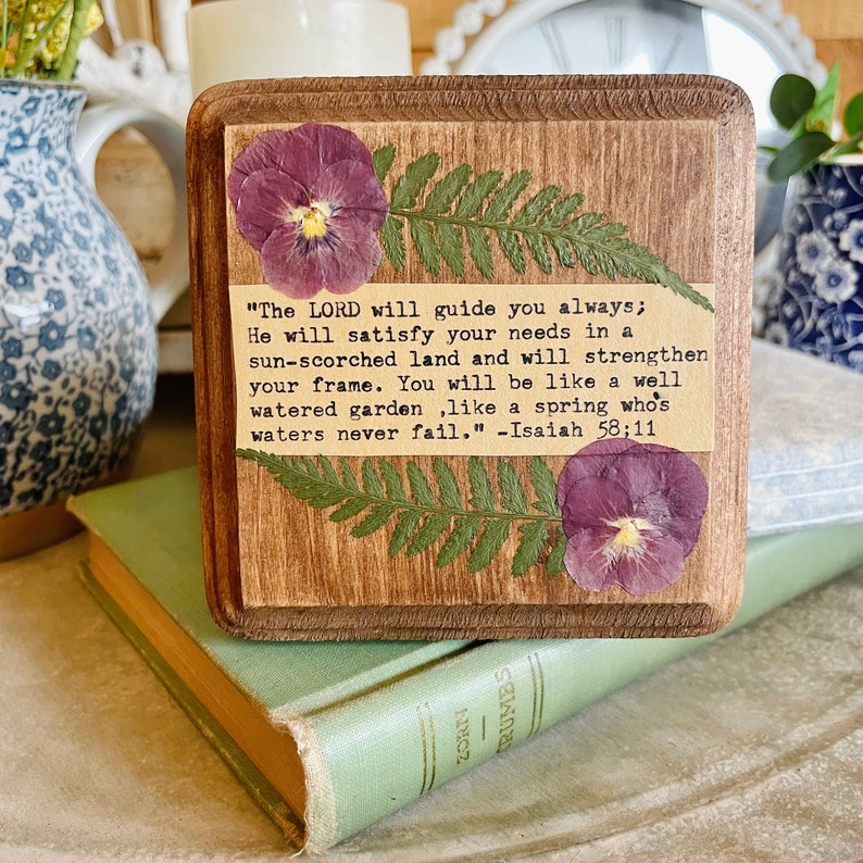 Op de afbeelding: Een houten bord met een ontwerp van geperste bloemen en een bijbelvers uit Jesaja 58:11. De tekst luidt: "De HEERE zal u altijd leiden; Hij zal in een door de zon verschroeid land in uw behoeften voorzien en uw kracht versterken. U zult zijn als een goed bewaterde tuin, als een bron waarvan de wateren nooit opdrogen."