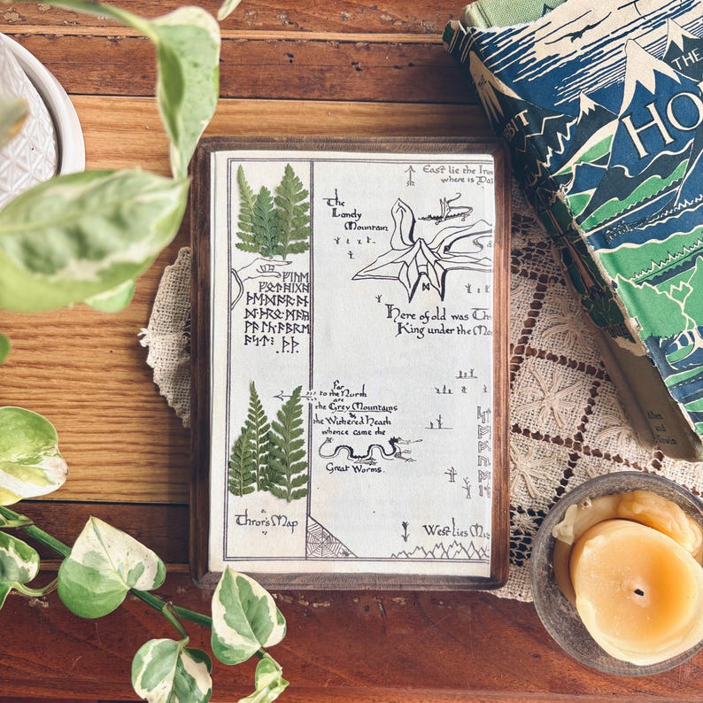 May include: A framed map with pressed ferns, featuring text and illustrations. The map is in a wooden frame, with a book titled "The Hobbit" visible in the background. A small bowl with candles and greenery are also present.