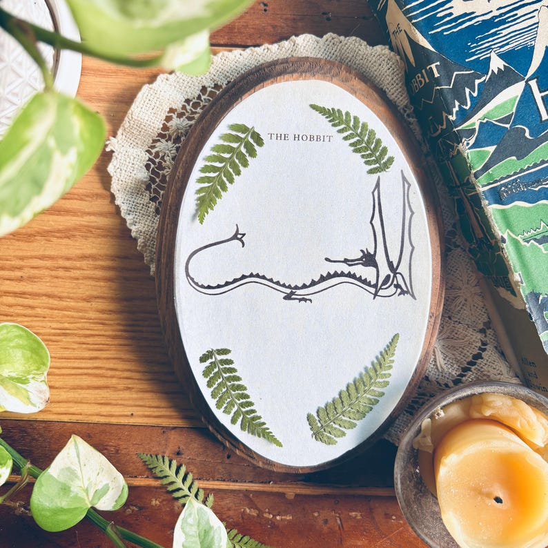 May include: Oval wooden plaque with the text "THE HOBBIT" and a dragon illustration. The plaque is decorated with green ferns and sits on a wooden surface. A book with a blue cover is visible in the background.