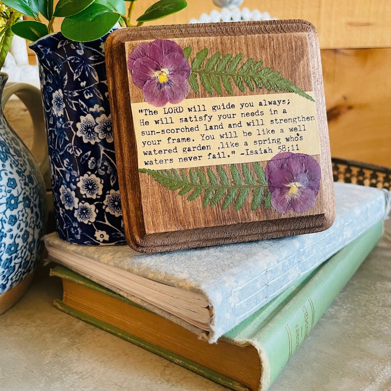 Op de afbeelding: Een houten bord met een ontwerp van geperste bloemen en een citaat uit Jesaja 58:11. Het citaat luidt: "De HEERE zal u altijd leiden; Hij zal in een dorre aarde in uw behoeften voorzien en uw kracht versterken. U zult zijn als een goed bewaterde tuin, als een bron waarvan de wateren nooit opdrogen." -Jesaja 58:11