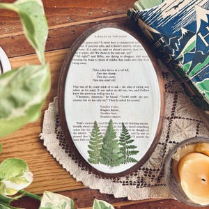May include: An oval wooden framed art piece with a fern design. The artwork features pressed ferns on a white background with text from "Riddles in the Dark" from The Hobbit. A vintage book is in the background.