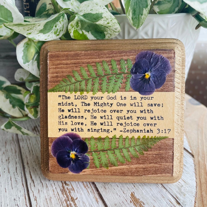 Op de afbeelding: Een houten bord met een rustieke bruine achtergrond en een beige rand. Het bord heeft een ontwerp met geperste bloemen met paarse viooltjes en groene varens. De tekst op het bord luidt: "De HEERE, uw God, is in uw midden, de Machtige zal u redden; Hij zal zich over u verheugen met blijdschap, Hij zal u met liefde stillen, Hij zal zich over u verheugen met zang." -Zefanja 3:17