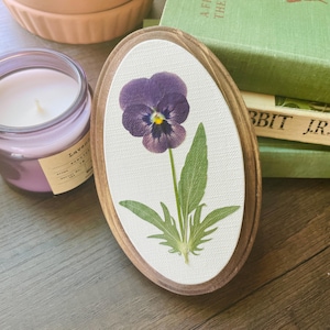 May include: Oval wooden frame with a pressed purple pansy flower on a white background. The flower has a yellow center and green leaves. A lavender candle and books are in the background.