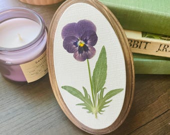 May include: Oval wooden frame with a pressed purple pansy flower on a white background. The flower has a yellow center and green leaves. A lavender candle and books are in the background.