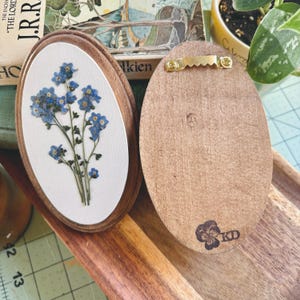 May include: Two oval wooden plaques. One plaque features pressed blue forget-me-not flowers on a white background within a dark wood frame. The other plaque shows the plain wood back with a gold-toned metal sawtooth hanger and a stamped logo.