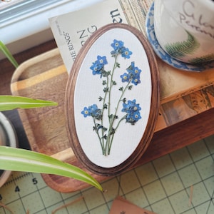 May include: An oval-shaped wooden frame containing a white background with pressed blue forget-me-not flowers. The flowers feature small, five-petaled blue blossoms with yellow centers and thin green stems arranged in a vertical botanical display.