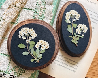 May include: Two wooden framed pressed flower art pieces. One is round, the other oval. Each features white flowers and green leaves on a dark blue background. The frames are a warm brown color. The pieces are displayed on a vintage book.