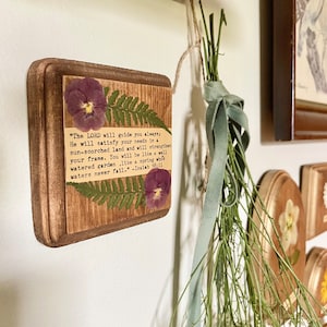 Op de afbeelding: Een houten plaquette met een ontwerp van geperste bloemen en een bijbelvers uit Jesaja 58:11. De tekst luidt: "De HEERE zal u altijd leiden; Hij zal in een dorre aarde in uw behoeften voorzien en uw kracht versterken. U zult zijn als een goed bewaterde tuin, als een bron waarvan de wateren nooit opdrogen." -Jesaja 58:11