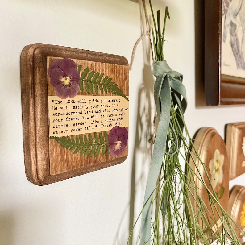 Op de afbeelding: Een houten plaquette met een ontwerp van geperste bloemen en een bijbelvers uit Jesaja 58:11. De tekst luidt: "De HEERE zal u altijd leiden; Hij zal in een dorre aarde in uw behoeften voorzien en uw kracht versterken. U zult zijn als een goed bewaterde tuin, als een bron waarvan de wateren nooit opdrogen." -Jesaja 58:11