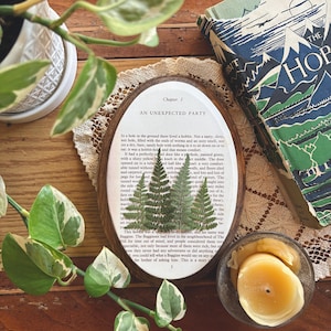 May include: Oval wooden framed art piece with pressed ferns on a page from "The Hobbit" book. The text reads "AN UNEXPECTED PARTY". A copy of the book and a small candle are also visible.