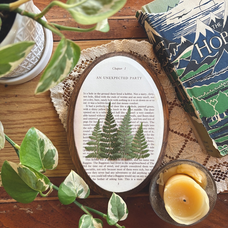 May include: Oval wooden framed art piece with pressed ferns on a page from "The Hobbit" book. The text reads "AN UNEXPECTED PARTY". A copy of the book and a small candle are also visible.