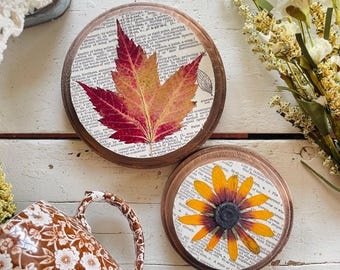 May include: Two round wooden coasters. One coaster features a pressed red and orange leaf, the other a yellow and brown flower. Both are set on a background of vintage book pages.