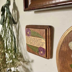 Op de afbeelding: Een houten plaquette met een ontwerp van geperste bloemen en een citaat uit Jesaja 58:11. De quote luidt: "De HEERE zal u altijd leiden; Hij zal in een dorre land in uw behoeften voorzien en uw kracht versterken. U zult zijn als een goed bewaterde tuin, als een bron waarvan de wateren nooit opdrogen." -Jesaja 58:11