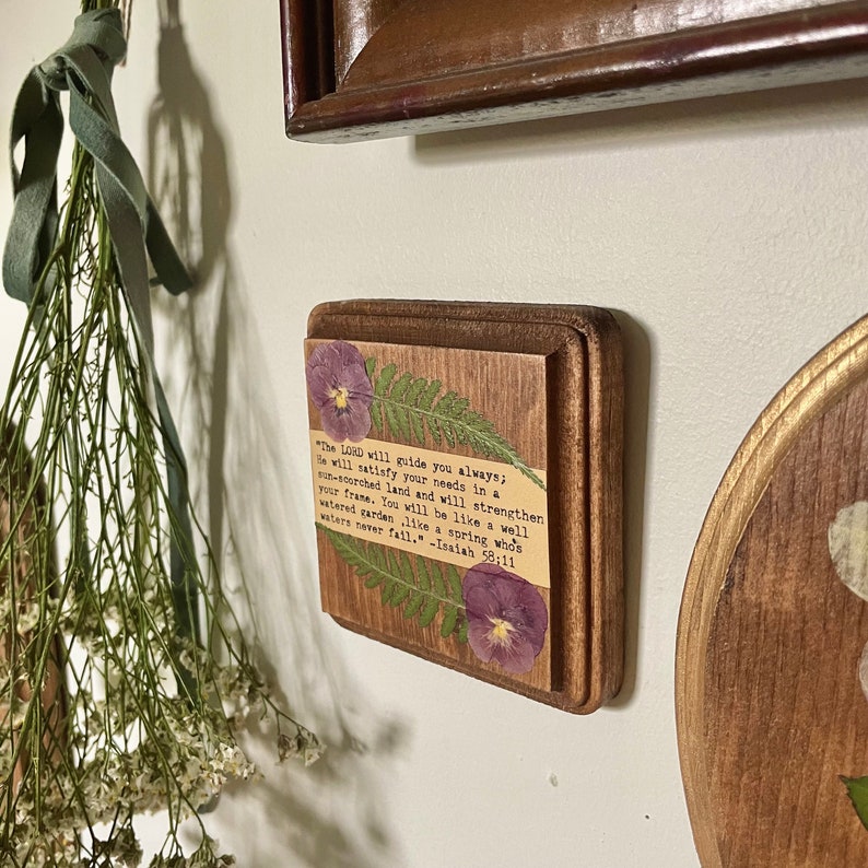 Op de afbeelding: Een houten plaquette met een ontwerp van geperste bloemen en een citaat uit Jesaja 58:11. De quote luidt: "De HEERE zal u altijd leiden; Hij zal in een dorre land in uw behoeften voorzien en uw kracht versterken. U zult zijn als een goed bewaterde tuin, als een bron waarvan de wateren nooit opdrogen." -Jesaja 58:11