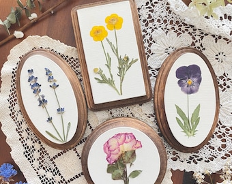 May include: Four oval-shaped wooden frames with pressed flowers inside. The frames are arranged on a white crocheted doily. The flowers include lavender, a purple pansy, a yellow buttercup, and a pink rose.