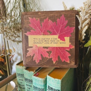 Puede incluir: Un letrero de madera con una cita de Ana de las Tejas Verdes sobre la belleza del otoño. La cita dice: "Estoy tan contenta de vivir en un mundo donde hay Octobres." El letrero está decorado con hojas de arce rojas prensadas.