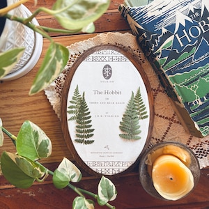 May include: An oval wooden frame with the book cover of "The Hobbit" by J.R.R. Tolkien. The book cover is white with green ferns and text. A vintage copy of the book is visible in the background, along with a lit candle and greenery.